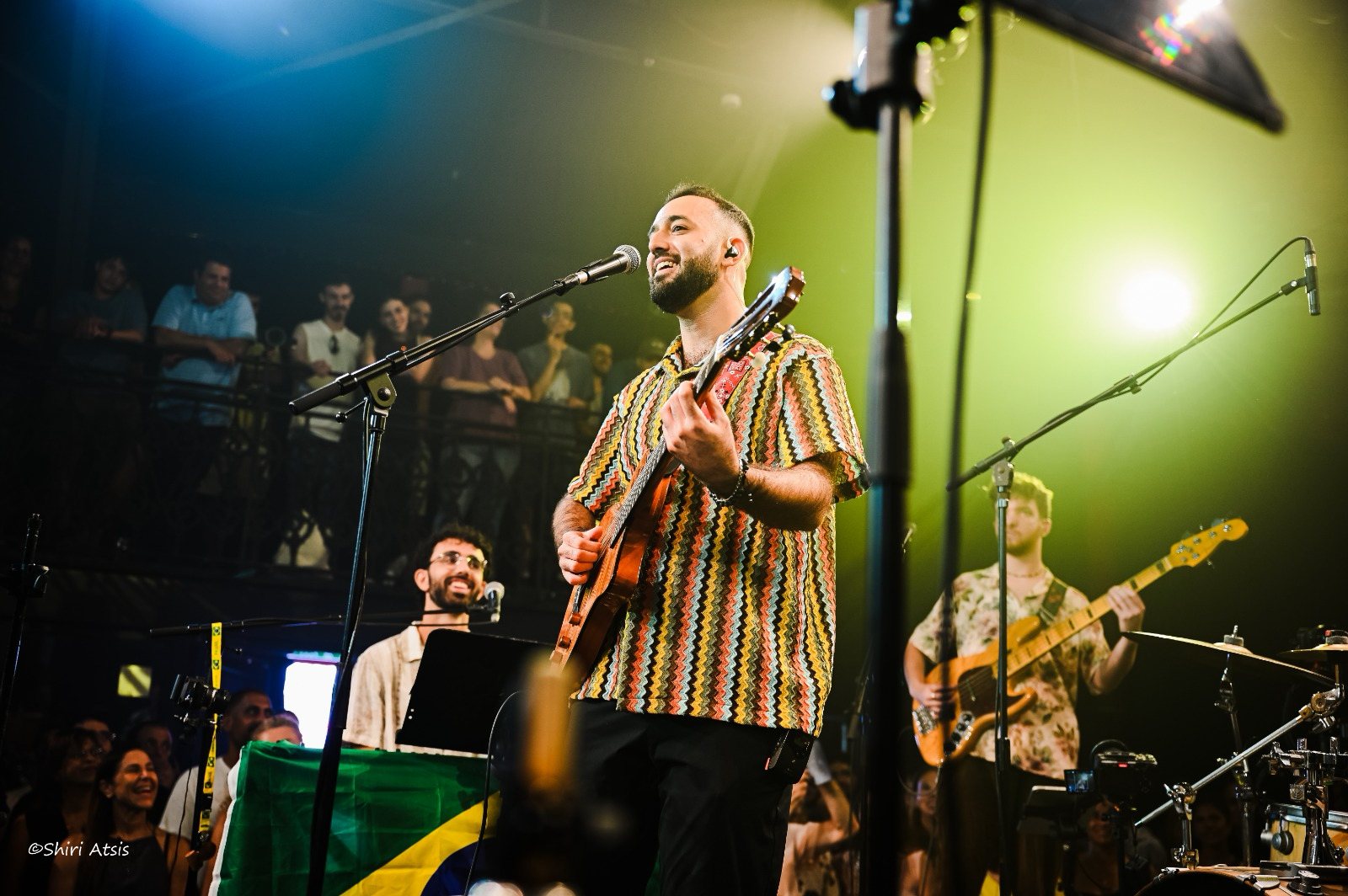 Amitai Kurtz on stage with guitar, a Brazilian flag on the keyboard behind him